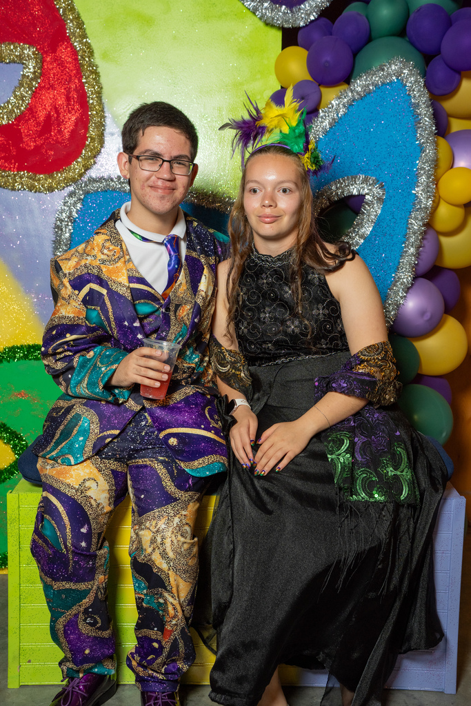 A couple dressed up in Mardi Gras smile together in front of a Mardi Gras backdrop inside the barn. 