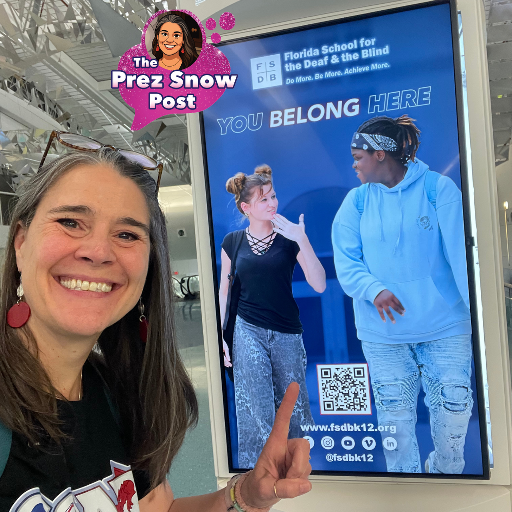 President Snow points at the digital display in the JAX airport showing two of our students walking and signing to each other with “You Belong Here” across the top with a QR code to our website.