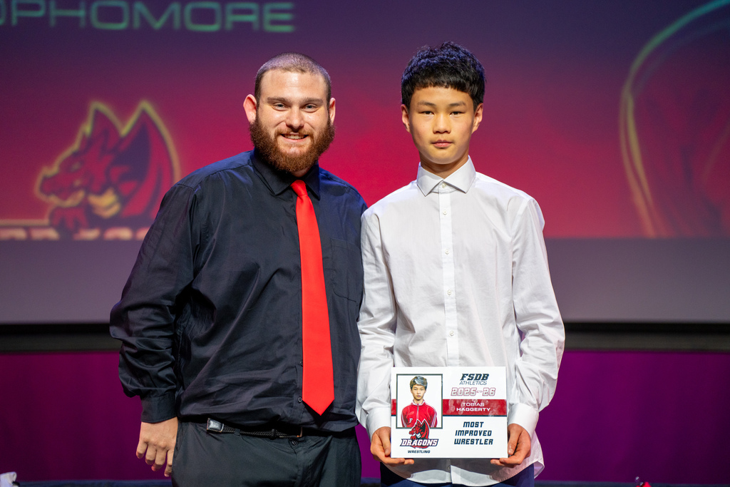 A student smiles on stage with a coach with his award.
