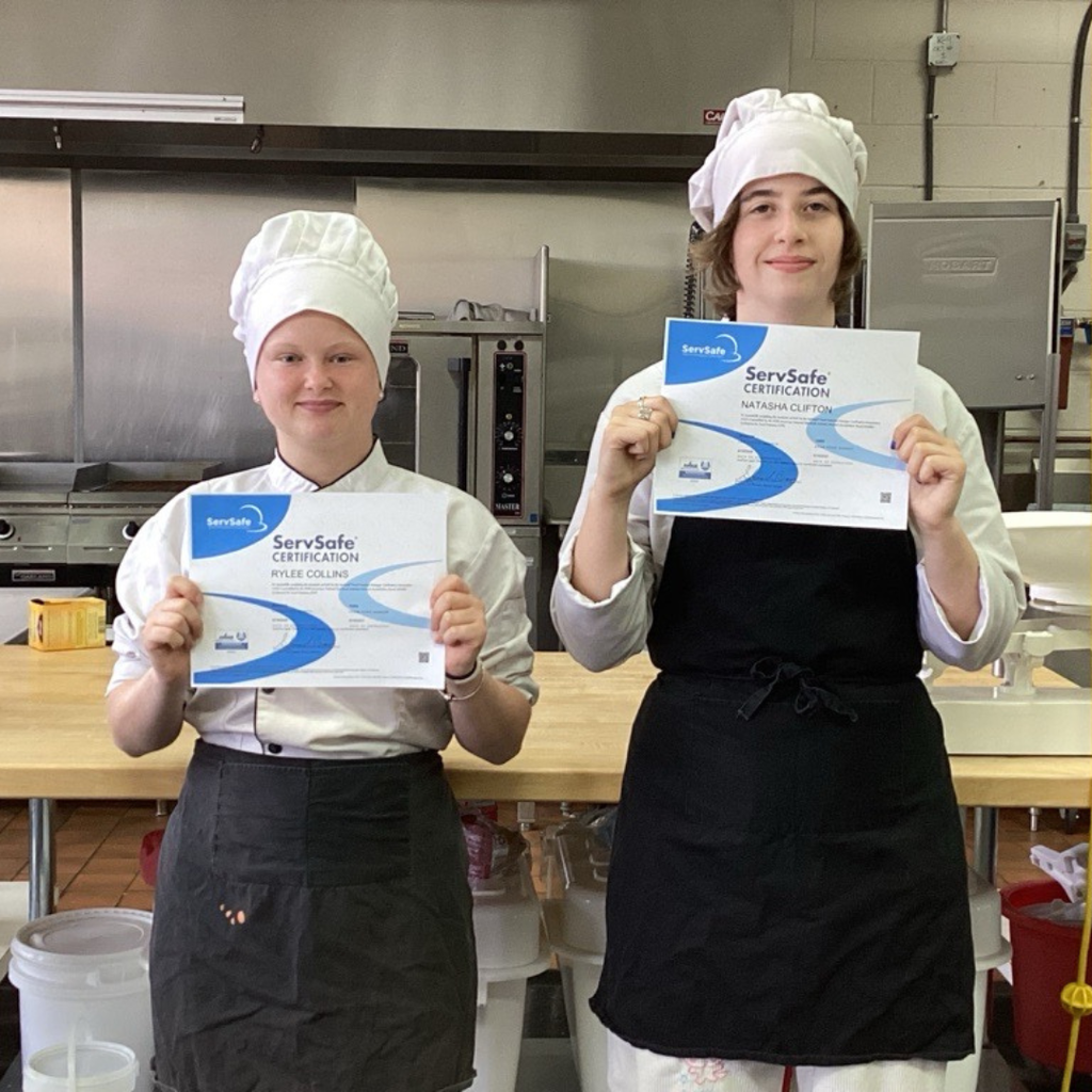 Two students dressed like chefs in white hat and black aprons smile with their certification inside the kitchen.