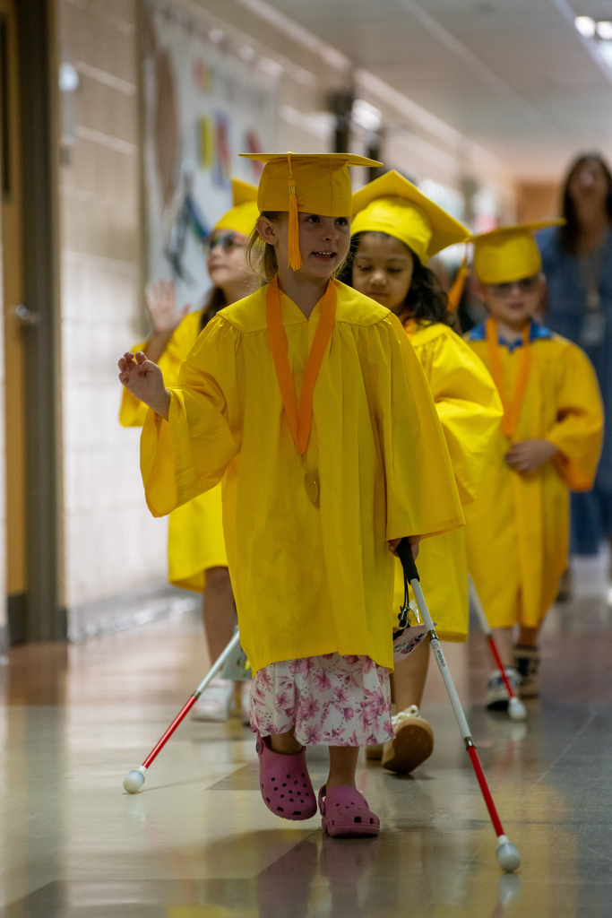 A Blind Kindergartener wearing yellow cap and gown wave as they walk down the hallway. 