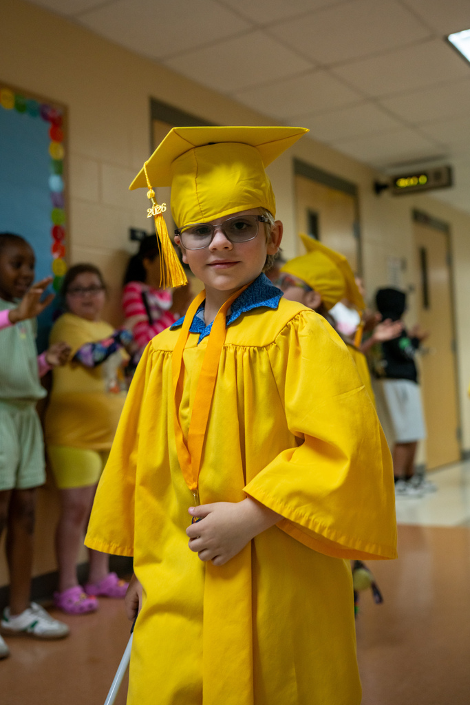 A Blind Kindergartener wearing yellow cap and gown wave as they walk down the hallway. 