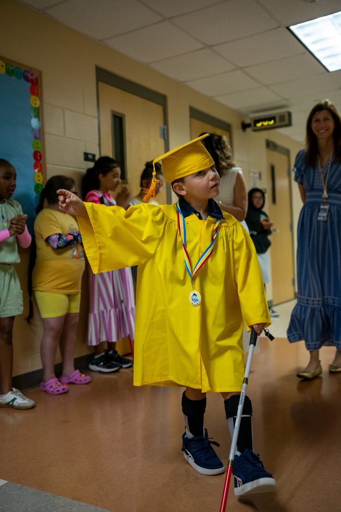 A Blind Kindergartener wearing yellow cap and gown wave as they walk down the hallway. 