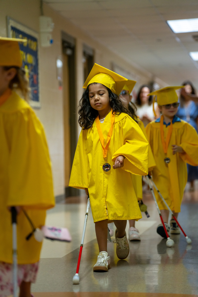 A Blind Kindergartener wearing yellow cap and gown wave as they walk down the hallway. 
