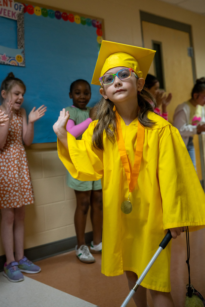A Blind Kindergartener wearing yellow cap and gown wave as they walk down the hallway. 