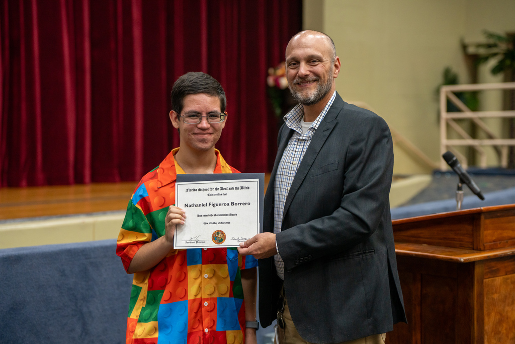 The Assistant Principal is holding an award that the student won smiling for the photo inside the Music Building. 