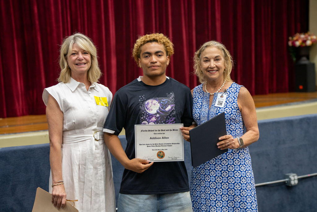 A student is holding his award and smiling with two ladies who gave him the award inside the Music Building. 
