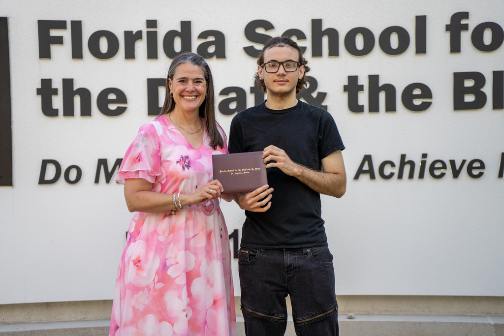 President Tracie Snow smiles with a student who holds his diploma by the FSDB sign. 