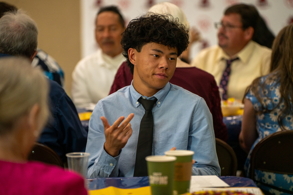 A senior wearing a blue shirt and black tie sign at the table he is sitting at. 