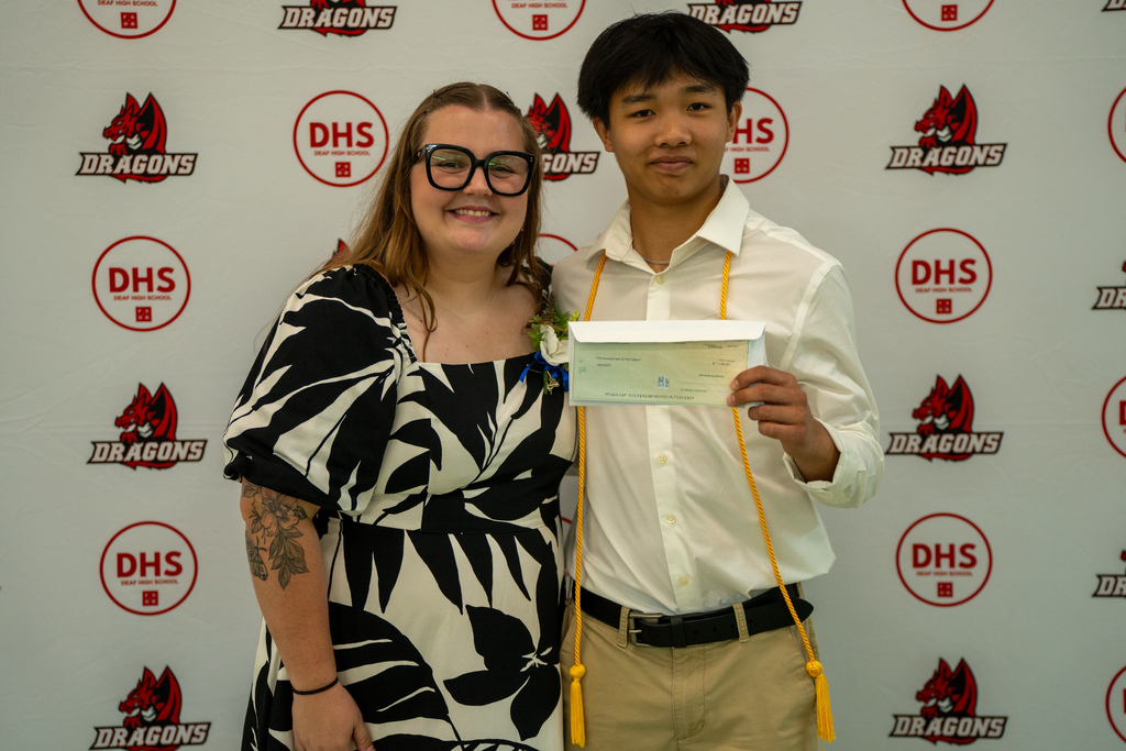 A senior smiles with a class sponsor holding a check in front of a DHS background. 