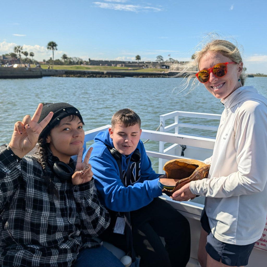 Two students pose with a female who works part of the group holding the turtle shell outside on the boat. 