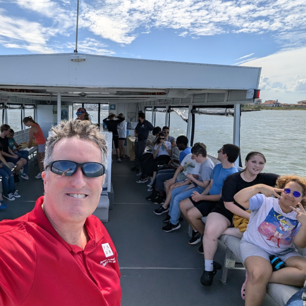 A selfie with the sponsor of the club and students in the back sitting on the boat. 