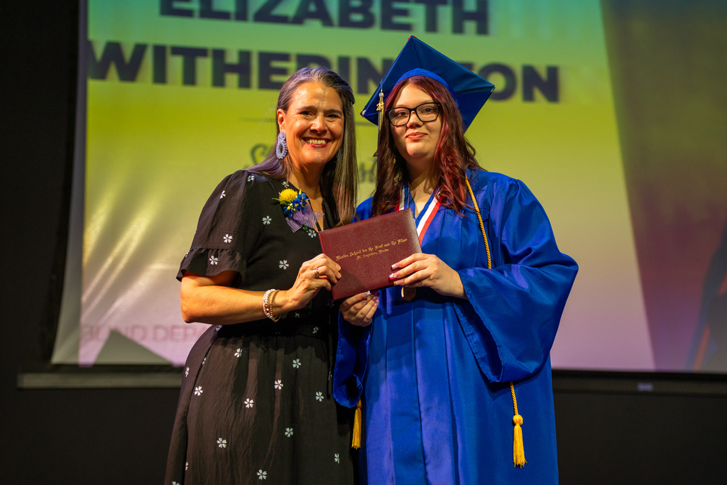 Tracie Snow smiles with a blind graduate holding the diploma on stage in Kirk. 