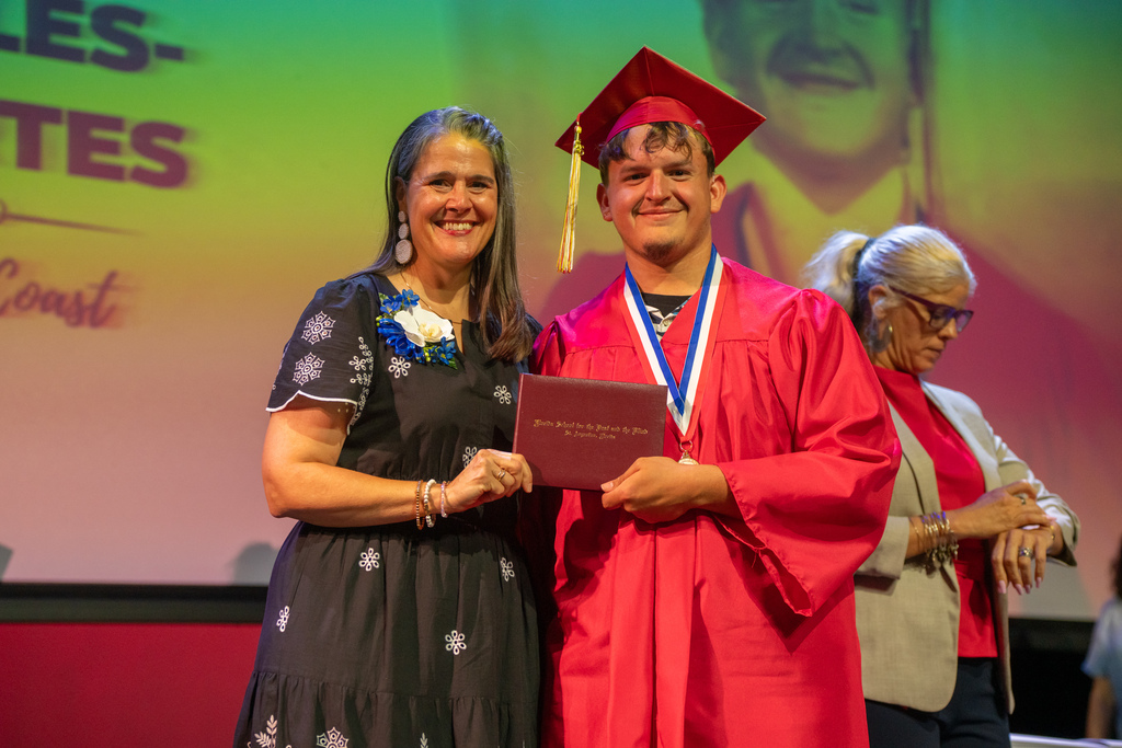 Tracie Snow smiling with a deaf graduate wearing red cap and gown on stage in Kirk. 