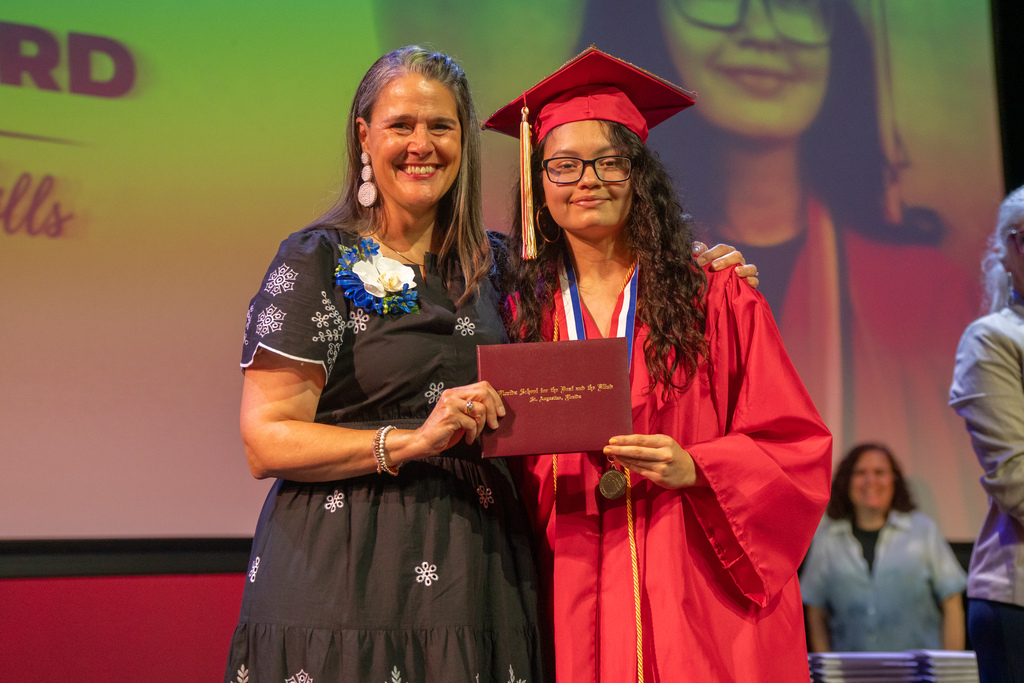 Tracie Snow smiles with a deaf graduate wearing red cap and gown on stage in Kirk. 
