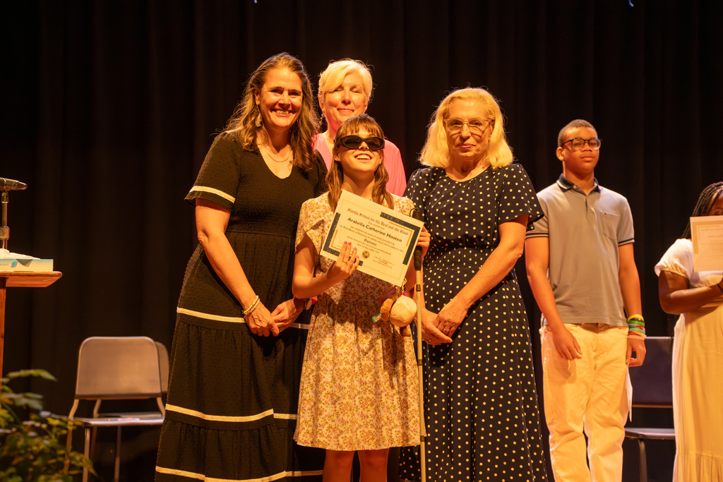 A group of staff smile with a 8th grade student who is holding her certification on stage in Music Building. 