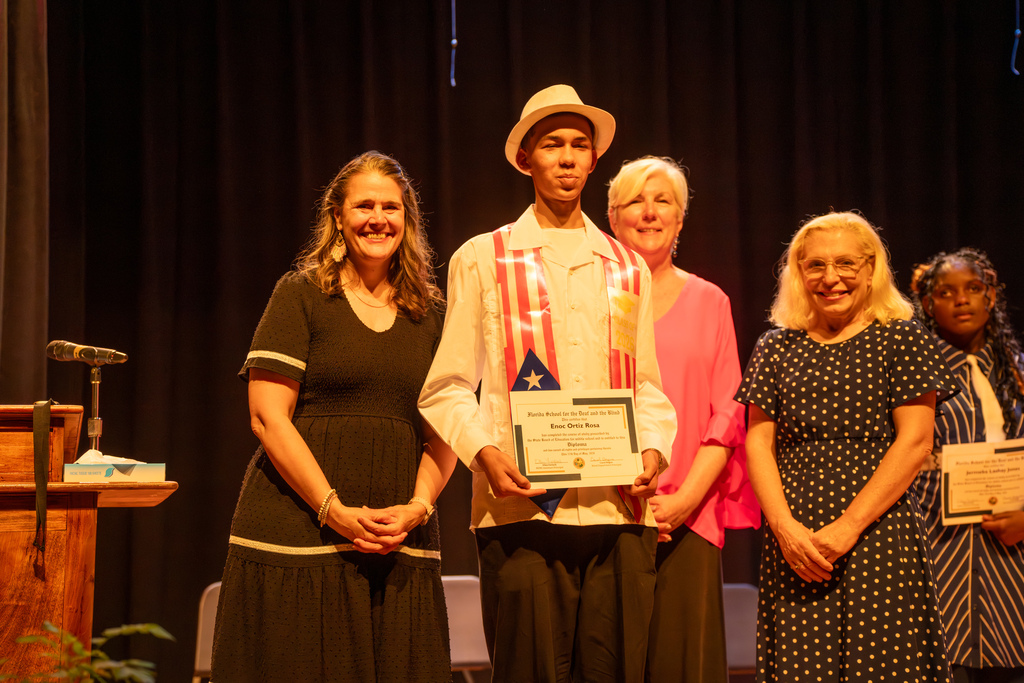 A group of staff members smile with 8th grade student who received his certification on stage in the Music Building. 