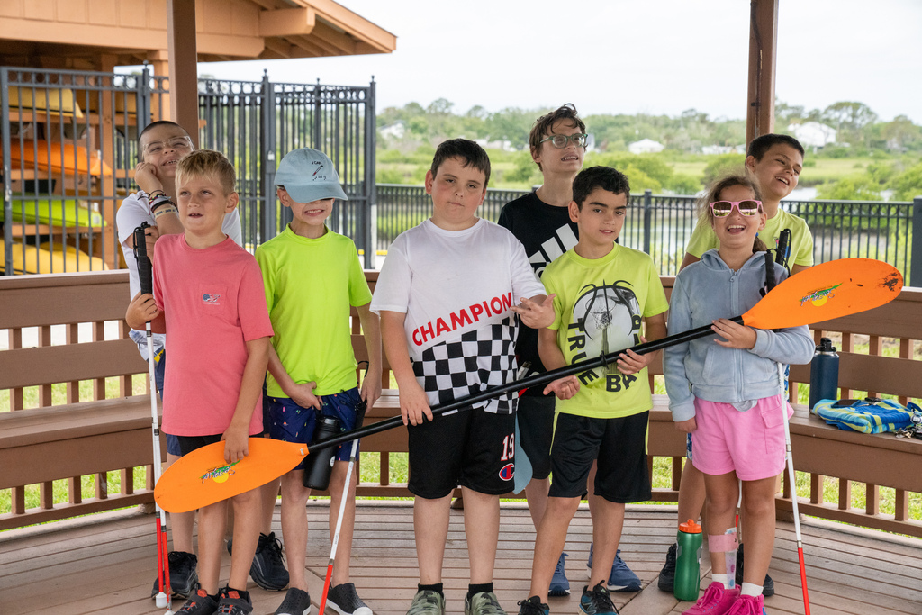 Group of students smile for a photo holding the kayak sticks under the Pavillon by the water at FSDB. 