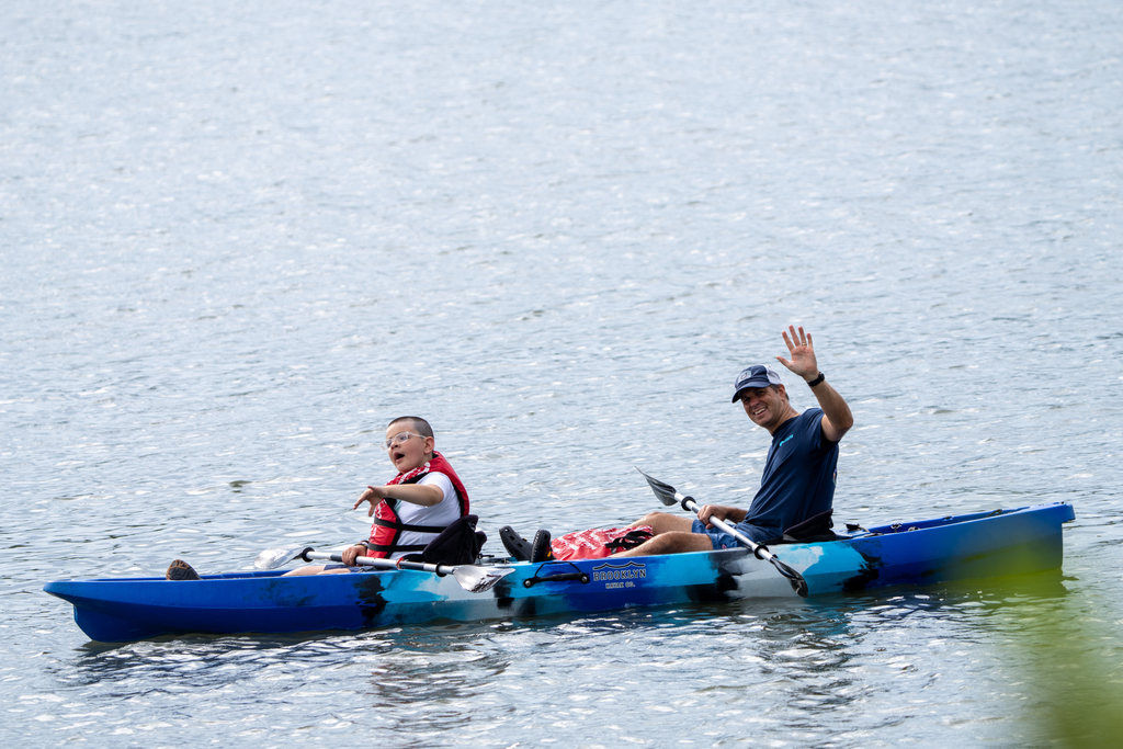 A camp staff member and camper wave to the camera out on the water in a blue kayak. 