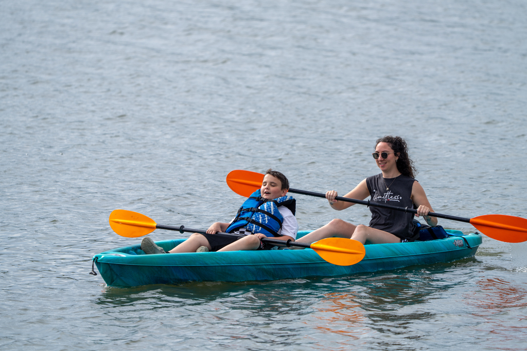 A camp staff member and a camper kayak in a blue kayak on the water. 