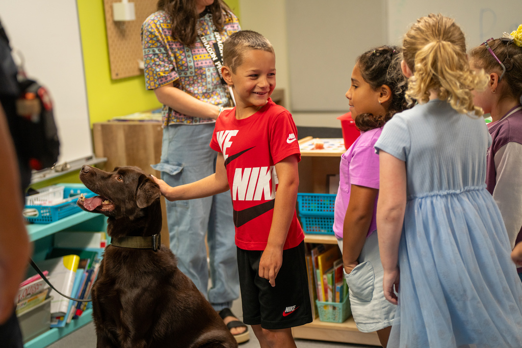 A student smiles as he pets the dog head inside the classroom.