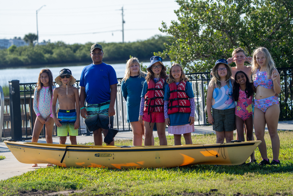 Summer Quest Group smile for a photo behind a yellow kayak by the water surrounding FSDB.