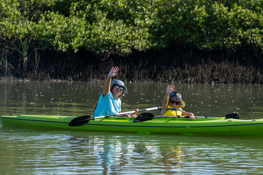 A camper and staff member sit in a green kayak out on the water and waving to the camera.
