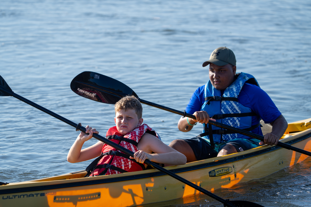 Two campers wearing life jackers and kayaking together in a yellow kayak out on the water.
