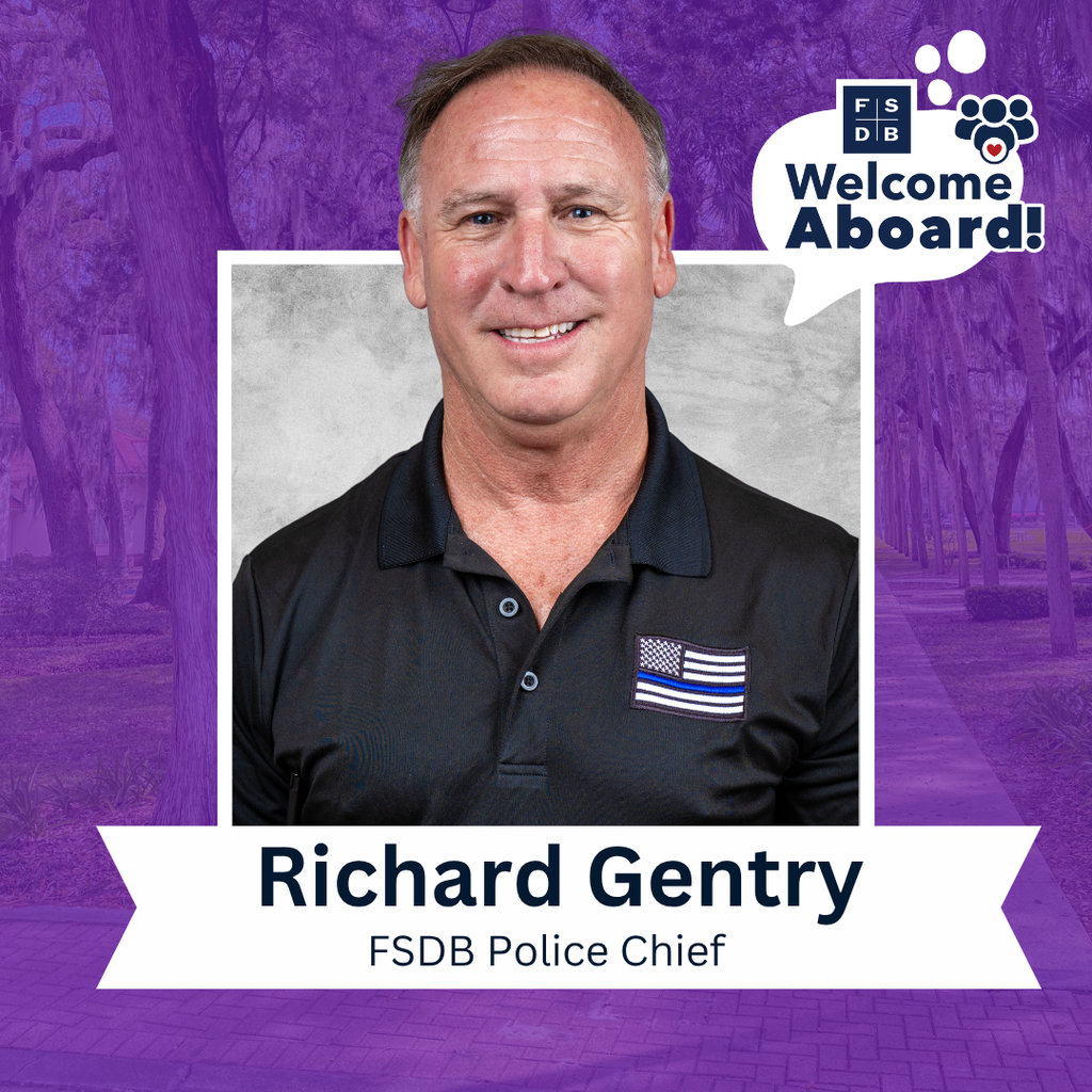 Richard is wearing a black collar shirt with American flag with blue line on his right chest smiles for a photo with a silver backdrop and purple background. Below the photo says "Richard Gentry, FSDB Police Chief" 