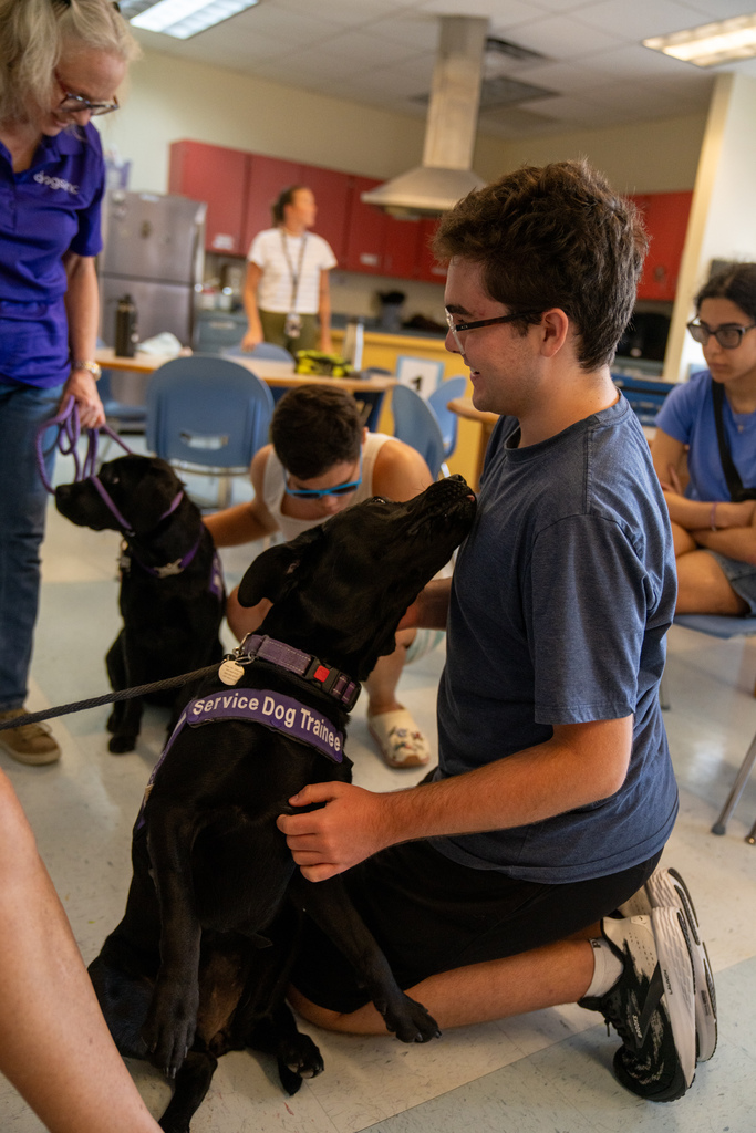 A camper smiles as the dog licks him inside the classroom. 