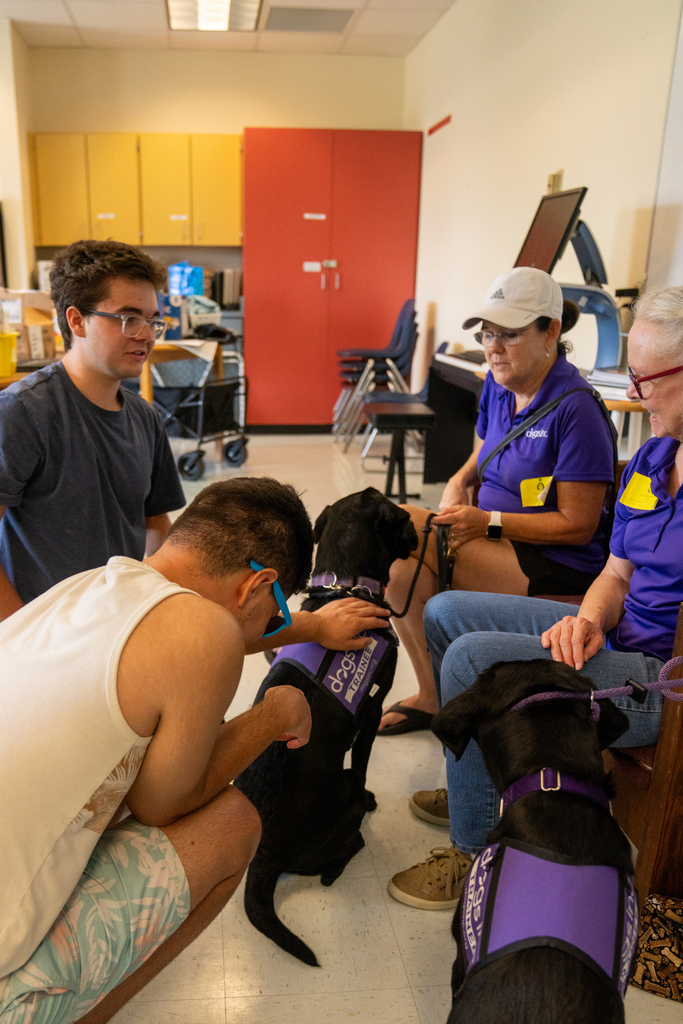 Two campers pet the black dogs while talking with two woman wearing purple shirts inside the classroom.