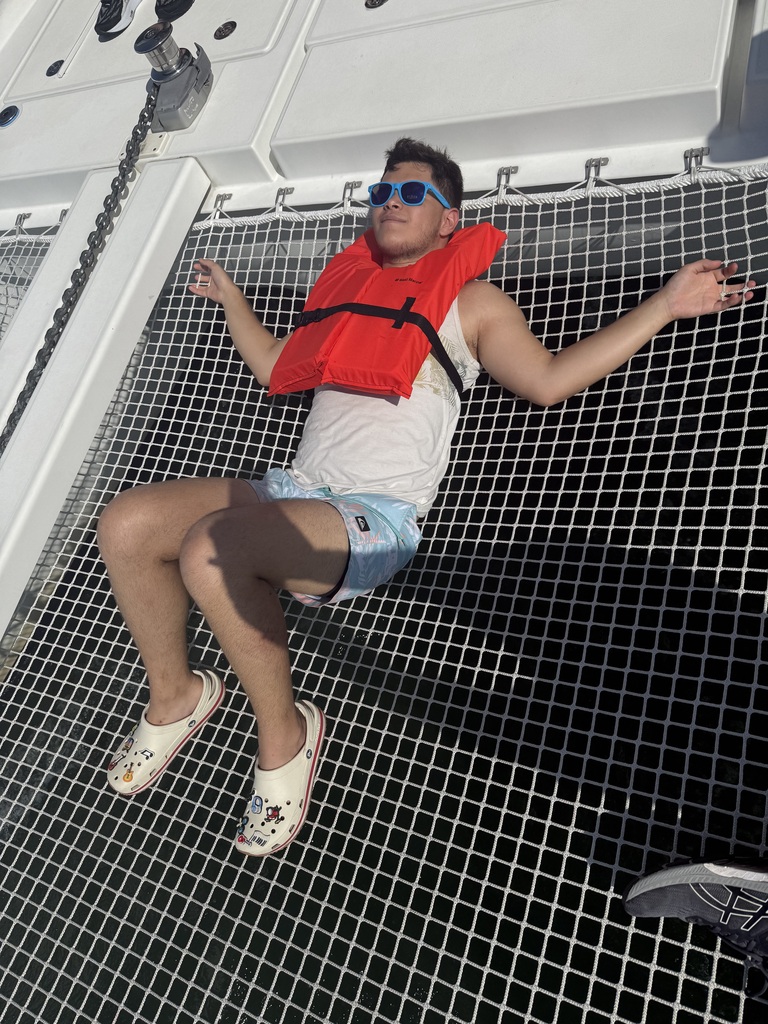 A camper lays down on the boat wearing a red life guard jacket and blue sunglasses. 