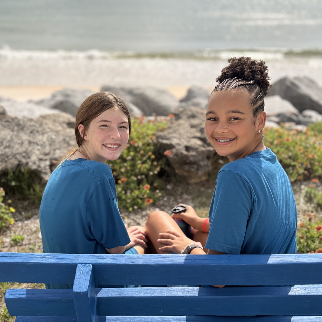 Two students sit on a blue chair and smile for a photo by the beach. 