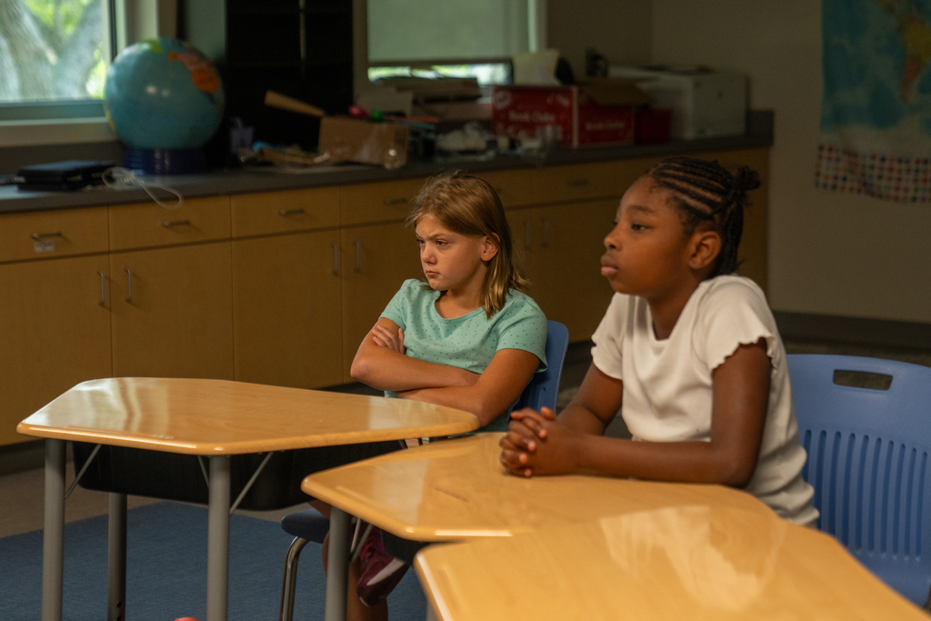 Two students sit at their desk listening to the teacher inside the classroom. 
