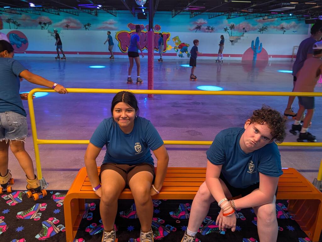 Two campers sit on a bench and pose for a photo inside the skating rink. 