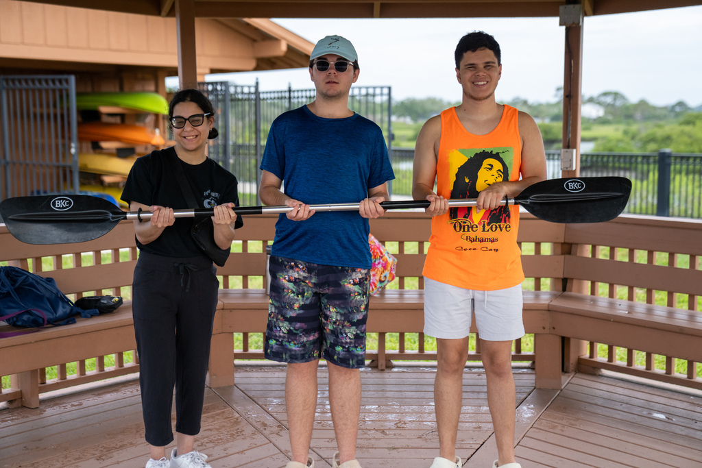 Three campers smile for a photo holding the kayak pole outside the Pavillon by the water.