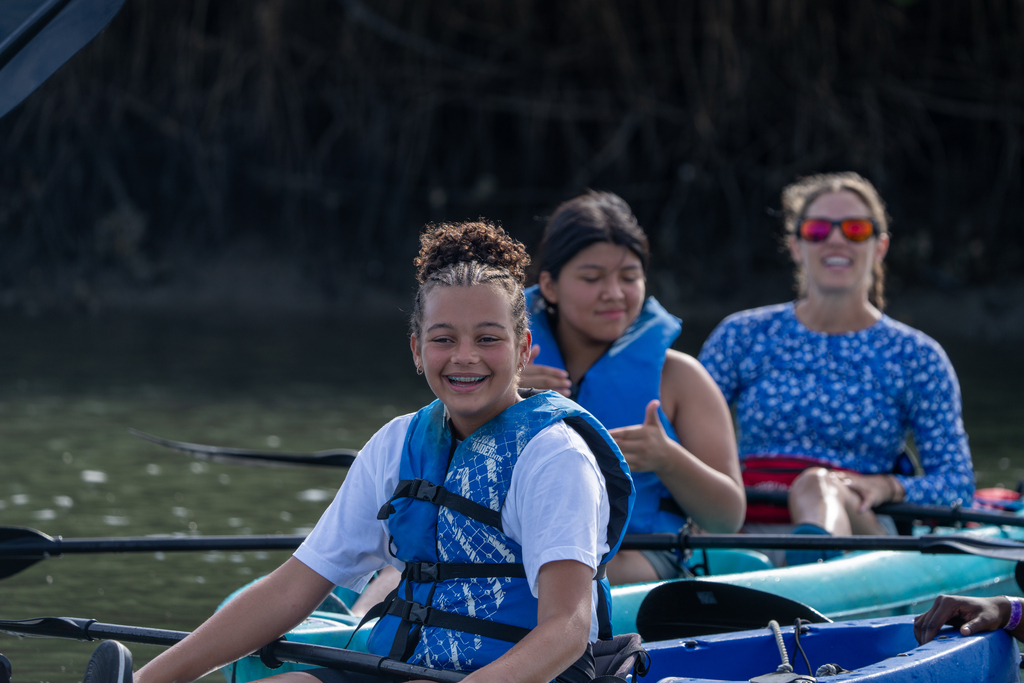 A camper wearing a blue life jacket vest and sitting in a blue kayak smile out on the water. 