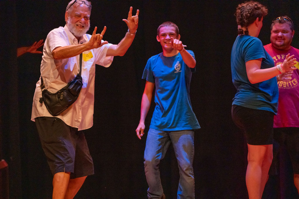 A camper points to the camera and his grandpa signs " I love you" on stage in the music building.