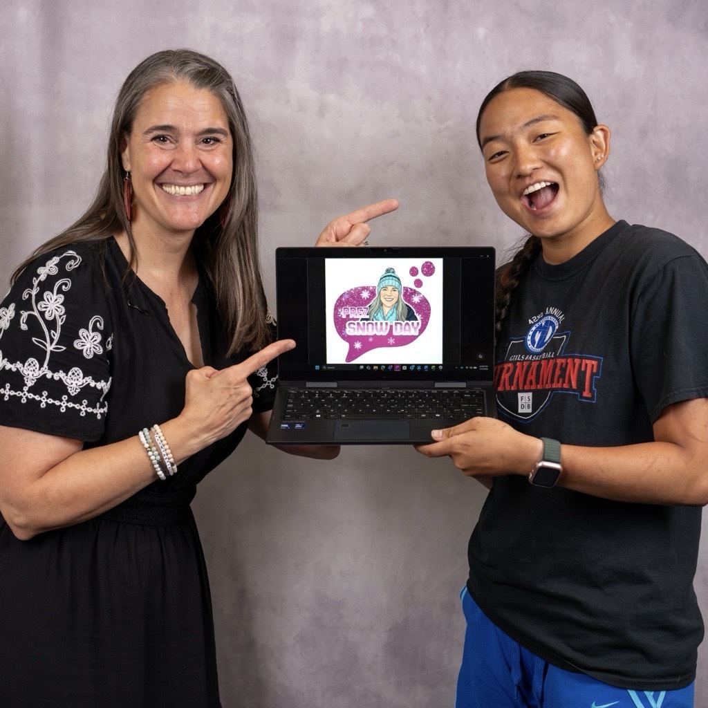 President Snow (left) and Emma Hancock (right) hold a laptop displaying the new "Prez SNOW Day" logo. President Snow is grinning pointing toward the screen and Emma holds the laptop with a huge smile. The laptop shows the new logo design which is a hot pink speech bubble with snowflakes, glitter, and a cartoon illustration of President Snow wearing a teal winter hat and scarf. The photo was taken against a neutral studio gray backdrop.
