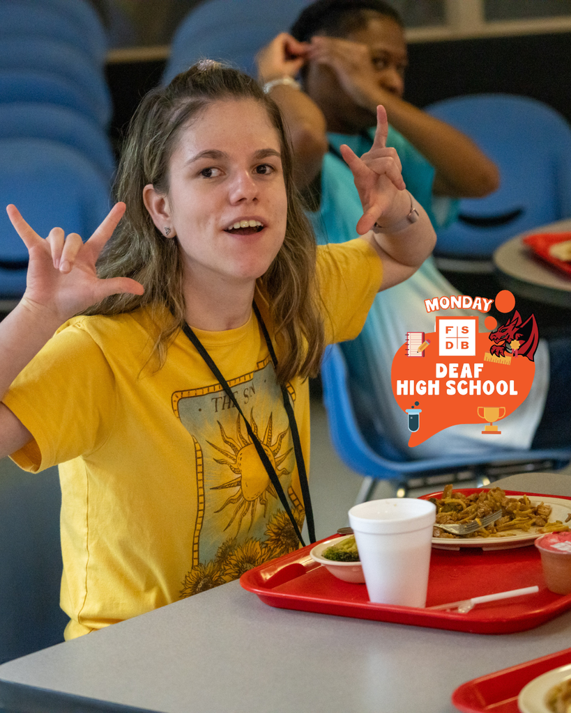 A student wearing a yellow shirt signs "ILY" for the photo while sitting down eating her lunch in DL.