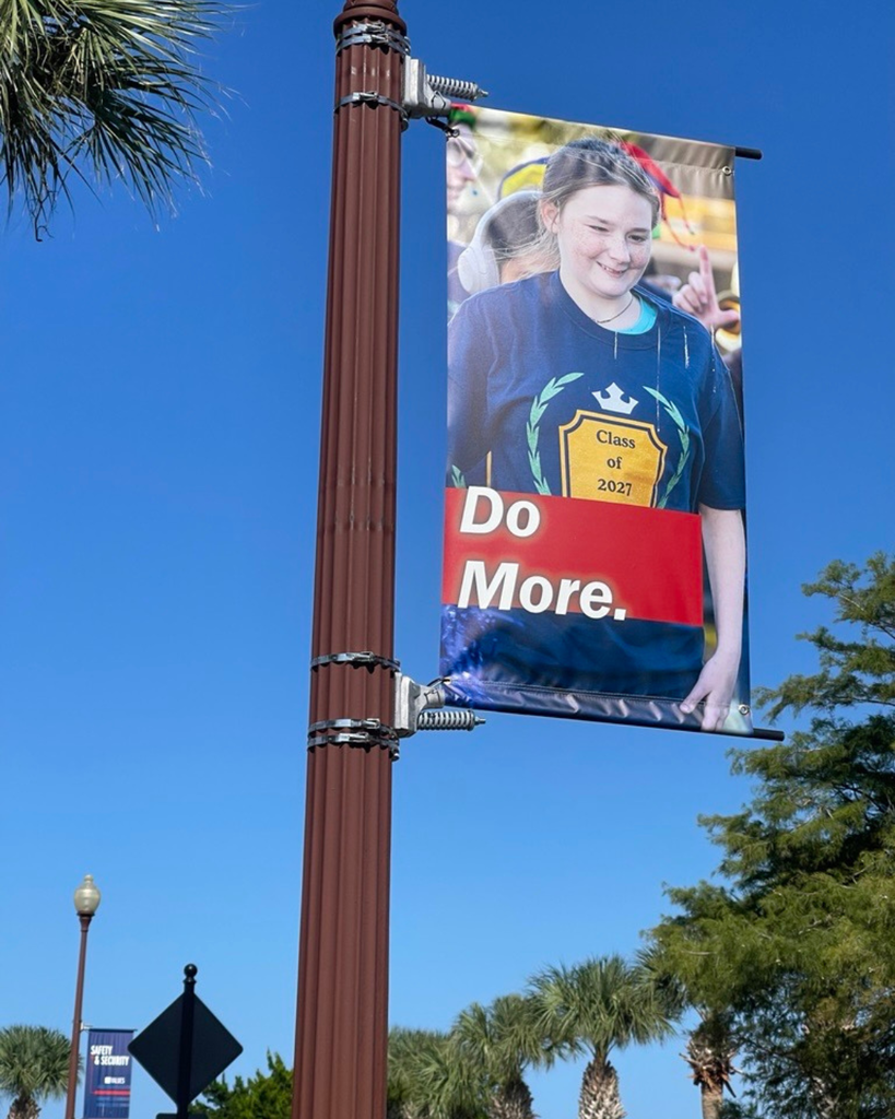 A light pole banner featuring a female student from the Blind High School wearing a shirt that says “Class of 2027” and the words “Do More.” The banner hangs against a clear blue sky, with palm trees visible in the background.