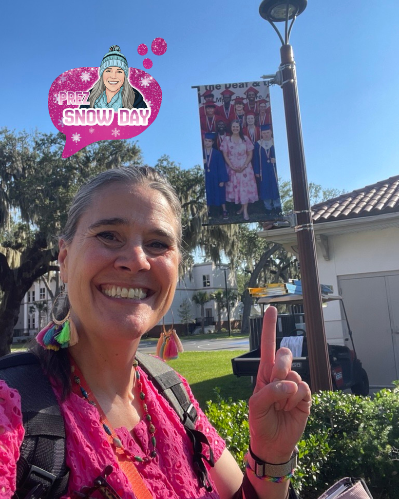 President Snow stands on the FSDB campus pointing toward a pole banner featuring a class photo of graduating students with her. The vertical banner is attached to a light pole with campus police building, trees, and a blue sky visible behind it.