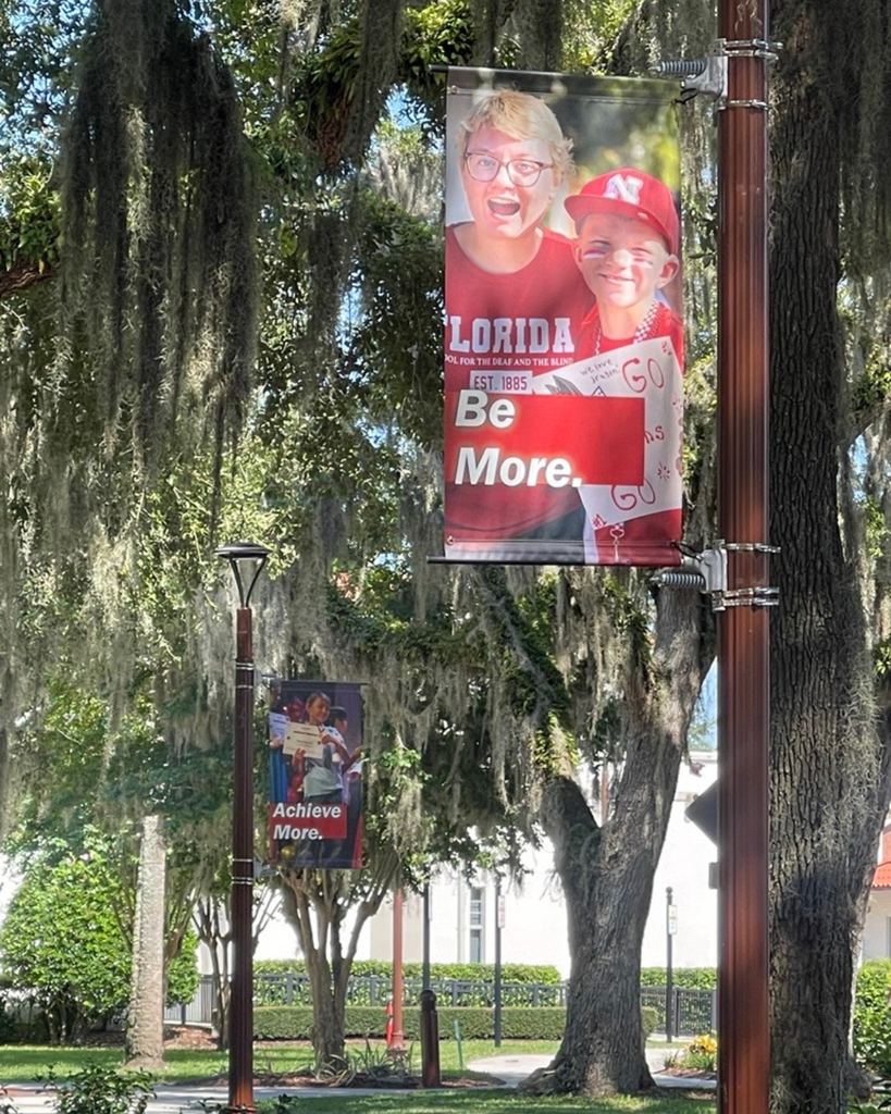 A campus banner displaying the words “Be More.” is mounted on a light pole beneath large oak trees draped with Spanish moss. The banner features a staff member and student standing side by side all dressed in red, while additional campus banners and trees can be seen in the background.