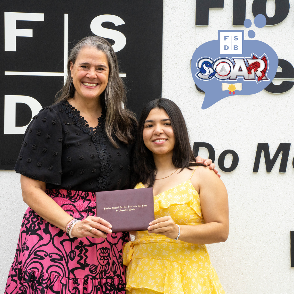 Tracie Snow is wearing a black and pink dress holding the diploma smiling with Andrea who is wearing a yellow dress smiles by the FSDB sign.