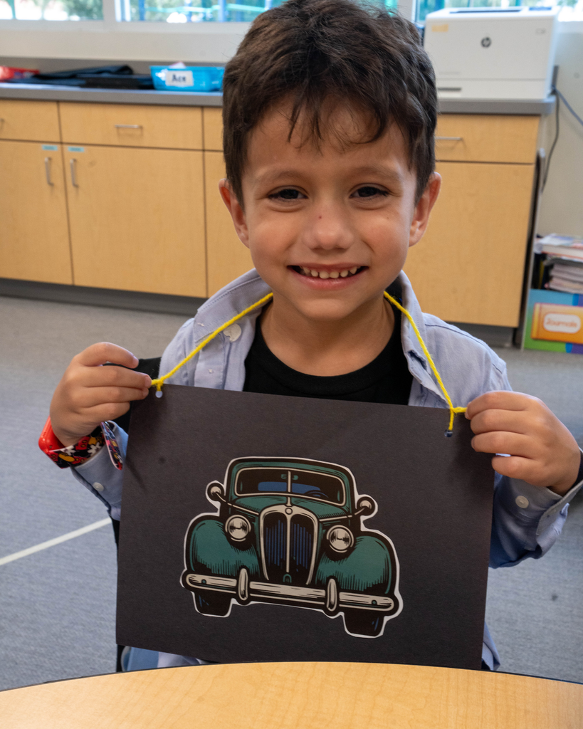 A student smiles with his car paper around his neck inside the classroom.