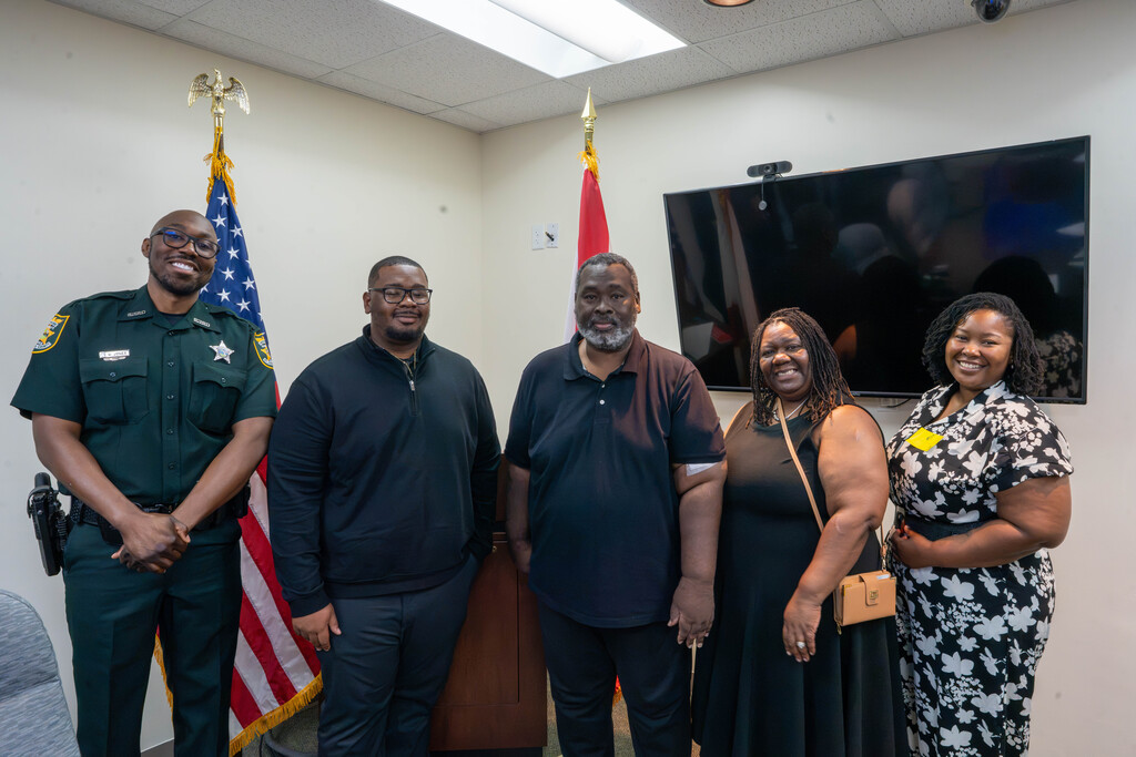 Ne'Darrius smiles with his family and a co worker who wears a green cop outfit inside campus police building .