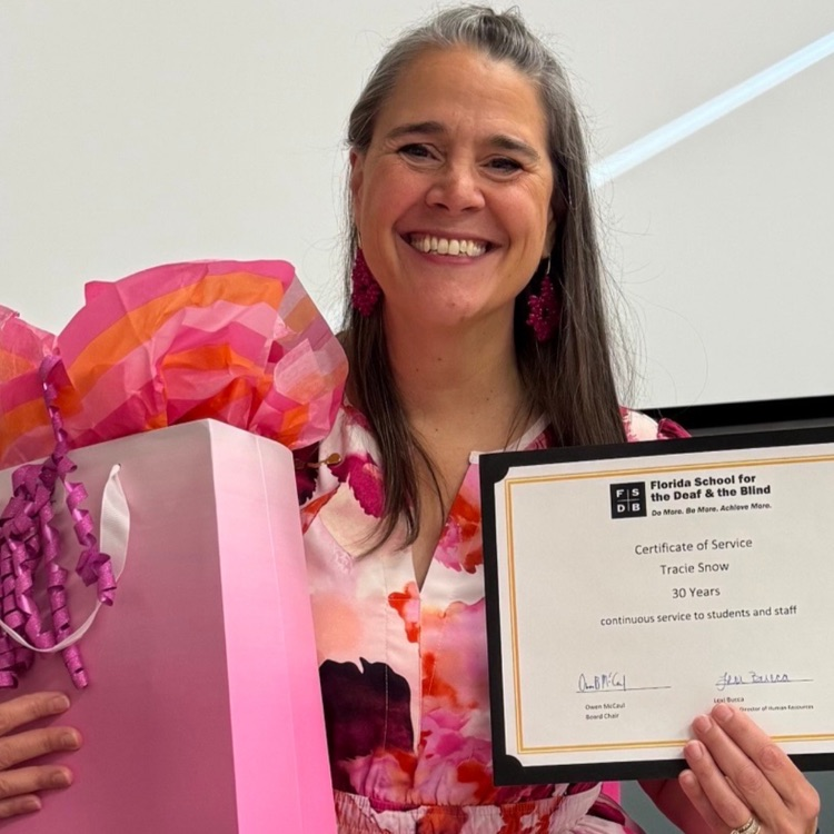 President Snow smiles brightly all dressed in pink while sitting at a table holding her certificate recognizing thirty years of continuous service at FSDB along with a gift bag presented in honor of the milestone. 