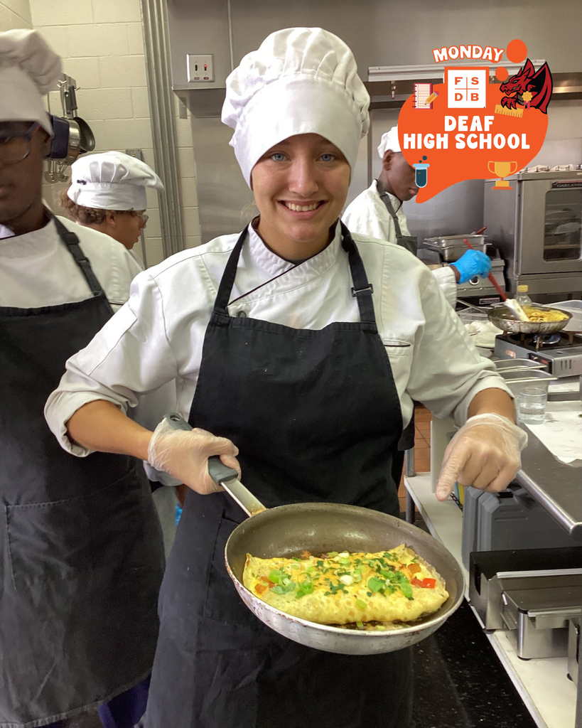 A student dressed in a chef hat and apron smile for a photo holding her omelet she made in a pan inside the kitchen. 