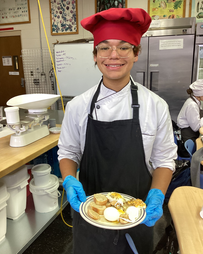 A student dressed in a chef hat and apron smile with his plate of eggs he created inside the kitchen. 