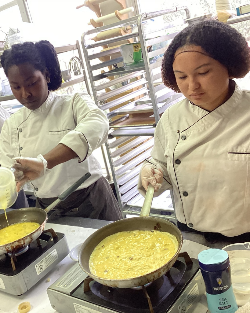 Two students dressed in  a chef hat and apron cook their eggs on a stove inside the kitchen. 